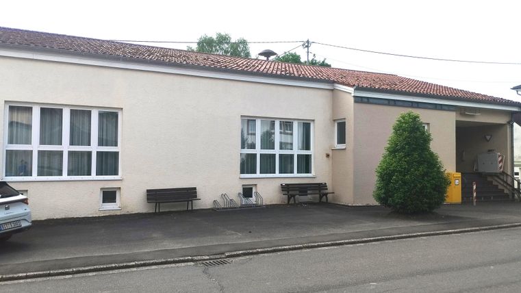 Un bâtiment simple avec une façade blanche et plusieurs fenêtres. Dehors, il y a des bancs et un petit arbre est visible.
