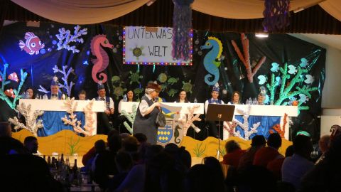 A festive event with a colorful backdrop of marine motifs. On stage, there are people in costumes, while one person is speaking.