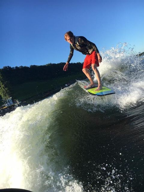Een surfer rijdt op een golf onder een heldere blauwe lucht. Op de achtergrond zijn groene heuvels zichtbaar.