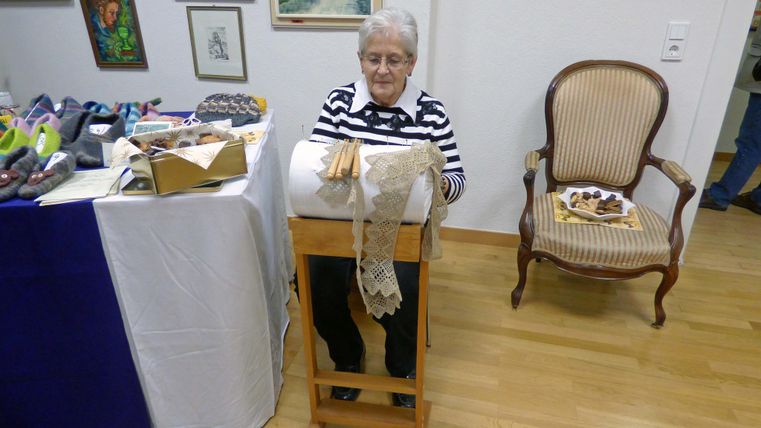 Une dame âgée est assise à une table et travaille sur un ouvrage de dentelle. À côté d'elle se trouve une chaise et quelques matériaux sur la table.