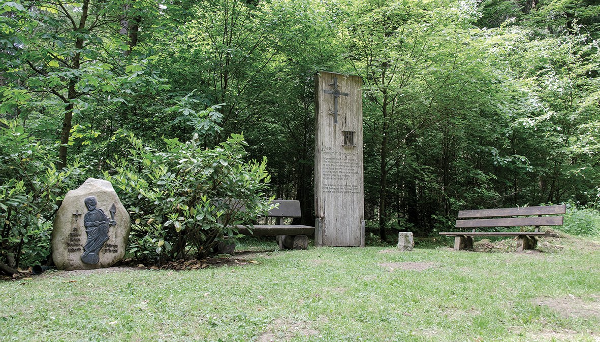 Ein Holzdenkmal und ein Steinrelief im Wald, umgeben von B&auml;nken und B&auml;umen. Die Szene wirkt ruhig und einladend., &copy; TI Bitburger Land