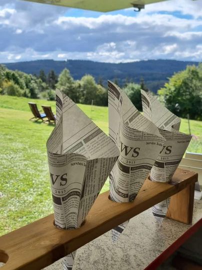 Three creatively folded paper containers made from newspapers are sitting on a wooden shelf. In the background, there is a picturesque landscape with green meadows and a cloudy sky.
