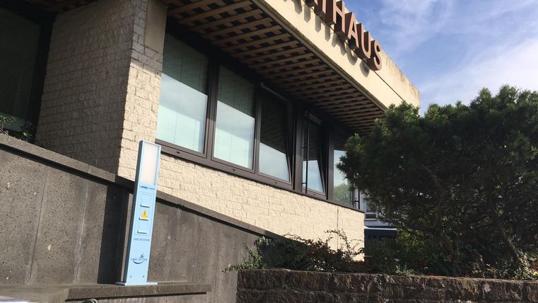 Un bâtiment moderne de mairie avec de grandes fenêtres et une enseigne. Au premier plan, on voit des buissons et un escalier.