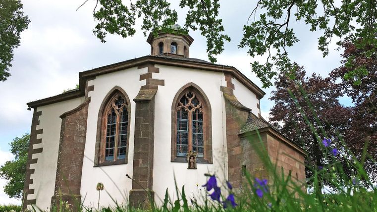 Eine kleine, charmante Kapelle mit gotischen Fenstern. Sie steht umgeben von grünem Gras und Bäumen unter einem bewölkten Himmel.