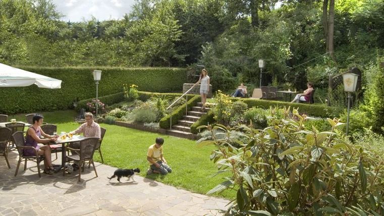 A beautiful garden with a lawn, blooming plants, and trees. People enjoy their time outdoors while a dog runs around.