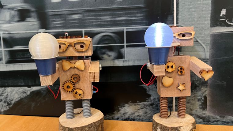 Zwei kreative Holzfiguren in Form von Robotern stehen auf einem Tisch. Jeder Roboter hält eine Lampe in der Hand und besteht aus verschiedenen Materialien und Formen.