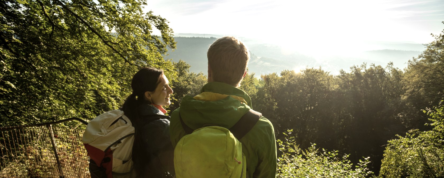 Zwei Wanderer mit Rucks&auml;cken stehen an einem Gel&auml;nder und blicken in die sonnige, bewaldete Landschaft., &copy; Dominik Ketz