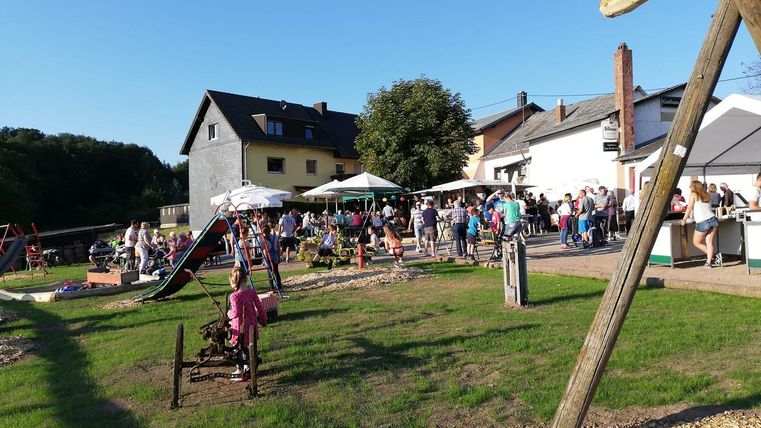 Ein belebtes Fest im Freien mit vielen Menschen, Ständen und Spielmöglichkeiten. Im Hintergrund sind Häuser und Bäume sichtbar.
