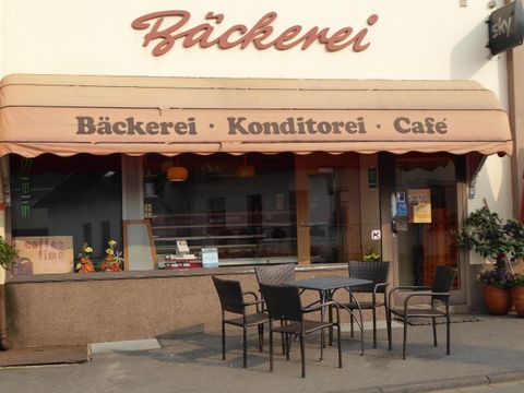 Eine gemütliche Bäckerei mit einem Café-Bereich und einem Sonnenschirm. Tische und Stühle stehen im Freien, um die Kunden willkommen zu heißen.