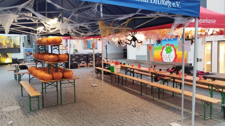 An event area with several tables and benches. In the middle, there are orange pumpkins and decorative spider webs are visible.
