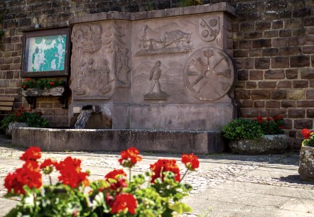 Ein Dorfbrunnen mit kunstvollen Reliefs aus Stein, umgeben von roten Blumen. Eine Infotafel und eine Bank sind ebenfalls sichtbar., &copy; TI Bitburger Land M. Mayer