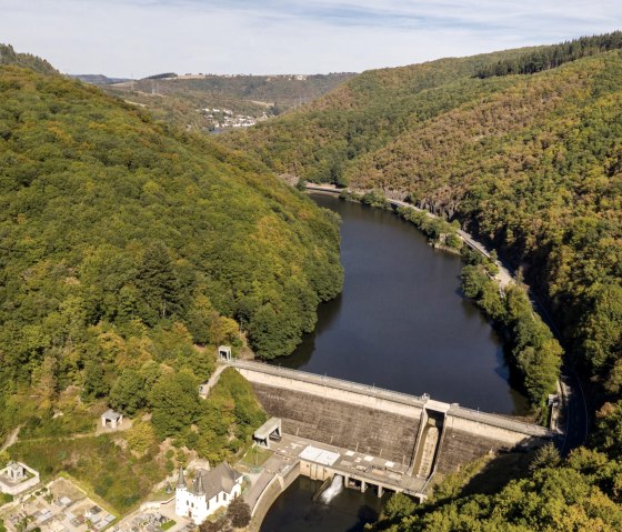 Vue a&eacute;rienne du lac de barrage de la vall&eacute;e de l'Our avec des collines bois&eacute;es et un mur de barrage. Au premier plan, on peut voir des b&acirc;timents et des chemins., &copy; Eifel Tourismus GmbH, D. Ketz