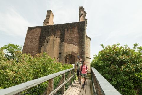 An old ruin surrounded by dense greenery. Two hikers are crossing a wooden bridge towards the ruin.