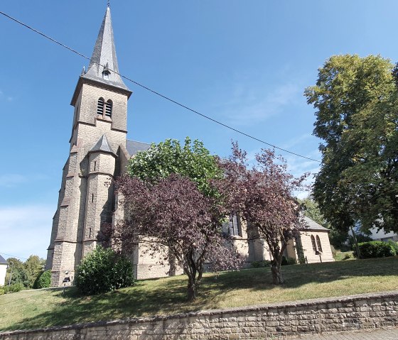 Die Pfarrkirche St. Bartholom&auml;us mit ihrem hohen, spitzen Turm steht umgeben von B&auml;umen unter einem klaren blauen Himmel., &copy; TI Bitburger Land - Steffi Wagner