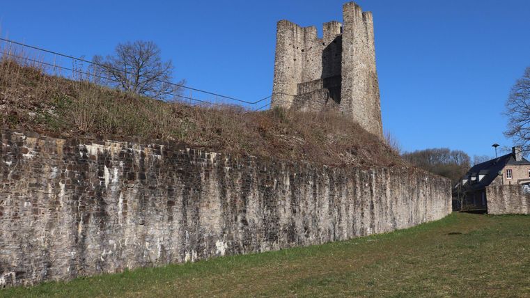 An old tower stands on a hill, surrounded by grass and trees. In the background, a clear blue sky can be seen.