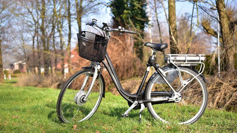 Un vélo noir avec un panier est posé sur une pelouse verte. À l'arrière-plan, on aperçoit des arbres et un ciel clair.