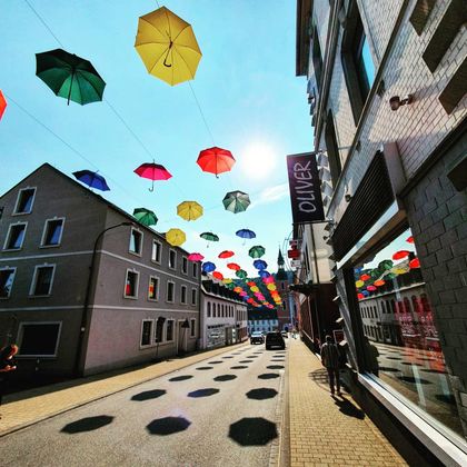 Eine Straße mit bunten Regenschirmen, die über den Bürgersteig hängen. Die Sonne scheint und wirft Schatten auf den Boden.