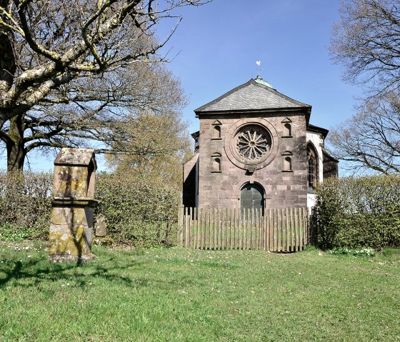 De Frohnertkapel in Oberkail, omringd door bomen en een oude grafsteen op de voorgrond, bij zonnig weer., &copy; TI Bitburger Land