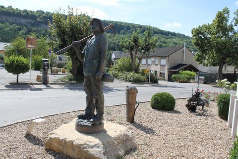 Une statue d'un homme avec un pic est située à un coin de rue. En arrière-plan, on peut voir des arbres et des maisons.