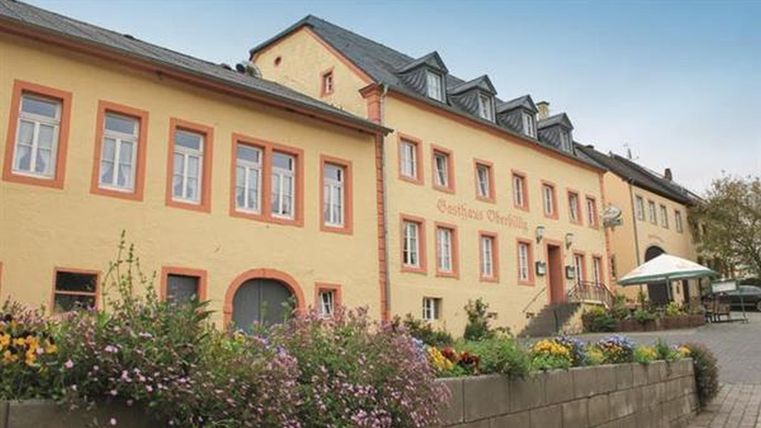 An elegant yellow building with several windows and a canopy. There are flower beds and a small square in the foreground.