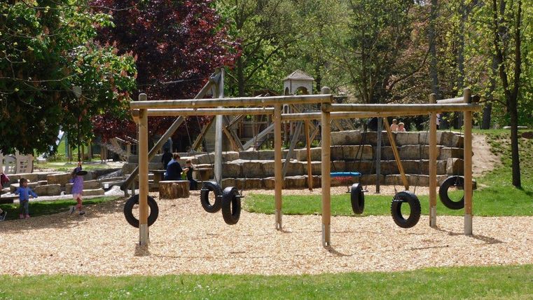 Ein Spielplatz mit Reifen-Schaukeln steht im Vordergrund. Im Hintergrund sind Baumhäuser und eine grüne Wiese zu sehen.