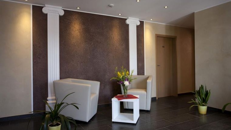A modern reception area with two bright seating areas and a small plant. Decorative columns are on the wall and a fresh flower arrangement is on a table.