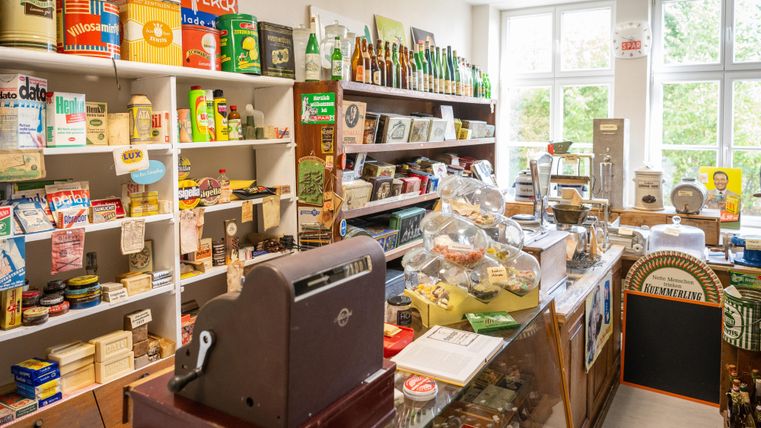 A cozy, nostalgic shop with shelves full of packaging and products from past times. In the foreground, there is an old cash register, and there are many different items to discover.