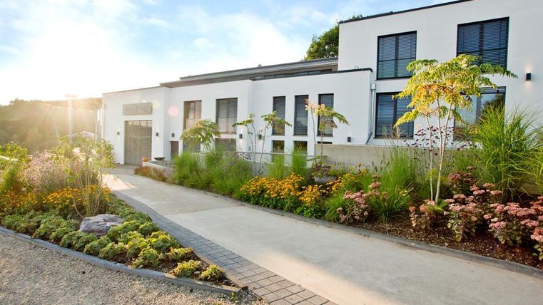 A modern building with large windows and an inviting entrance. The garden is lushly designed with colorful flowers and green plants.
