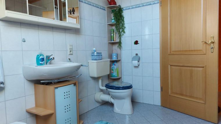 A bright bathroom with white tiles and a wooden cabinet. It features a sink, a toilet, and a blue rug.