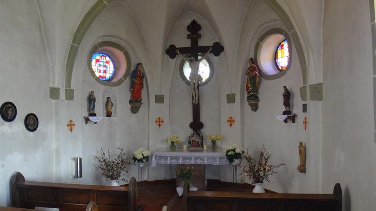 Eine kleine Kapelle mit einem Altar, gekreuzigten Jesus und bunten Fenstern. Bänke und religiöse Statuen sind ebenfalls sichtbar.