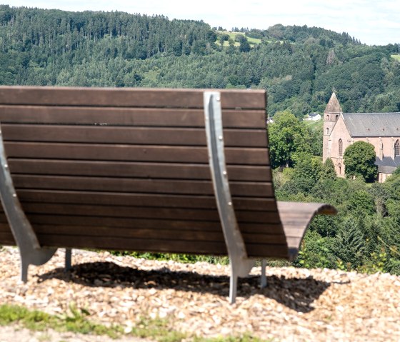 Een houten bankje op de voorgrond biedt uitzicht op een kerk omringd door groene bomen en heuvels.