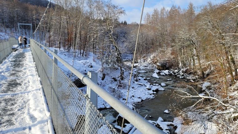 Eine schneebedeckte Hängebrücke über einen fließenden Bach. Umgeben von verschneiten Bäumen und einer klaren blauen Himmel.