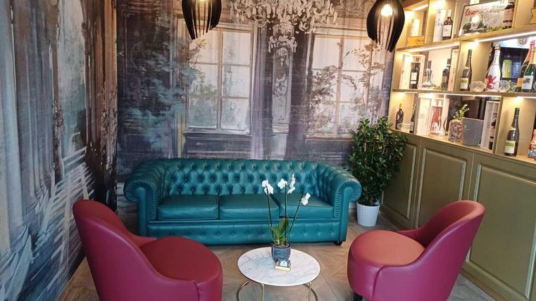 Een stijlvol ingerichte ruimte met een groene Chesterfield-bank en twee rode fauteuils. Aan de muur hangen decoratieve lampen en een plank met drankjes is zichtbaar.