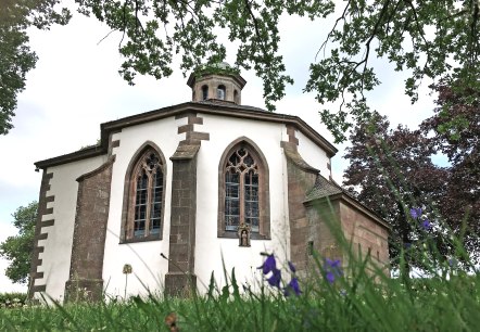 La chapelle Frohnert avec ses fen&ecirc;tres gothiques, entour&eacute;e d'arbres et de fleurs au premier plan, photographi&eacute;e depuis une perspective basse., &copy; TI Bitburger Land