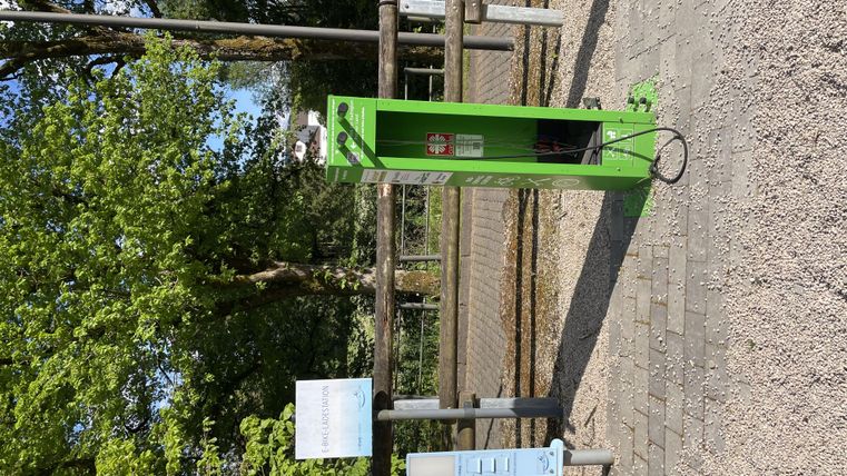 une station de charge verte pour VTT électriques se trouve sur un chemin entouré d'arbres. À côté de la station se trouve un panneau d'information.