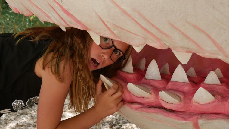 Ein Mädchen mit Brille posiert in einem kunstvollen Modell eines Hai-Mundes. Sie schaut überrascht aus dem Maul des Hais heraus.