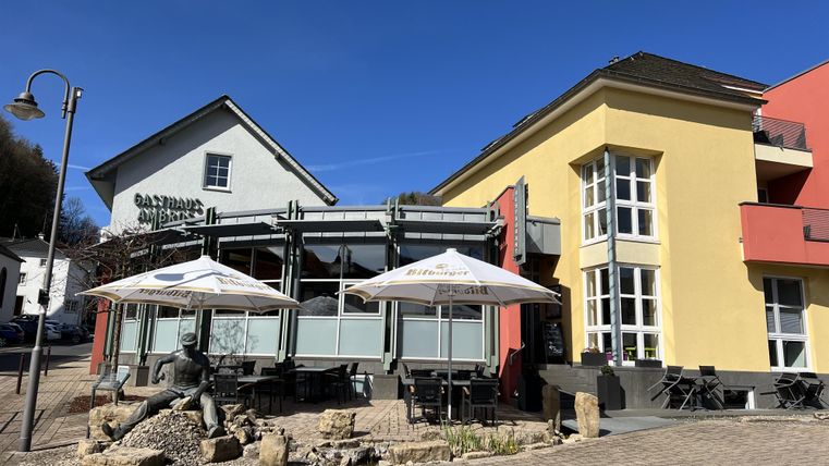 A terrace with parasols and seating in front of a restaurant. In the background, two buildings are visible, one in light yellow and the other in white.