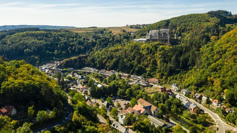Luftaufnahme von Vianden mit Schloss auf einem Hügel, umgeben von Wald und Häusern.