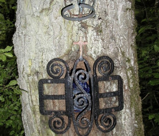 Un tronc d'arbre avec un ornement m&eacute;tallique artistique et une petite croix. L'ornement est encastr&eacute; dans l'arbre., &copy; Conny Meier