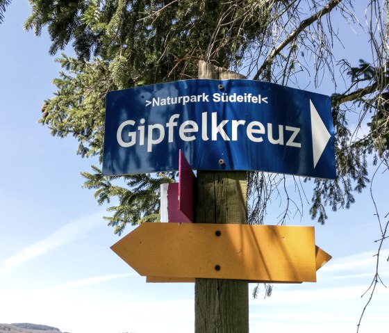 Ein Wegweiser im Naturpark S&uuml;deifel zeigt zum Gipfelkreuz. Der blaue Himmel und gr&uuml;ne Zweige rahmen das Schild ein., &copy; TI Bitburger Land