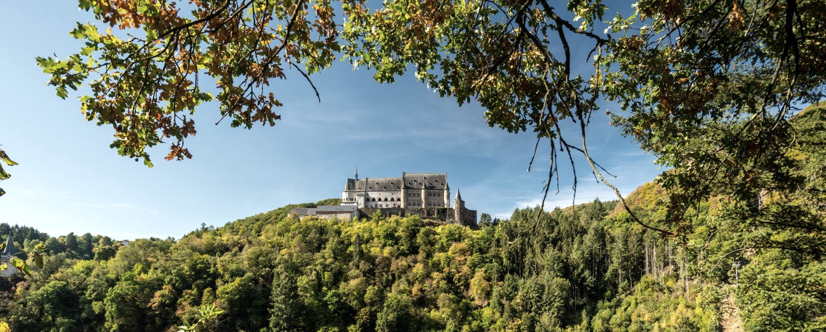 Le château de Vianden trône majestueusement sur une colline boisée, entouré d'arbres automnaux et d'un ciel bleu., © Eifel Tourismus GmbH, D. Ketz
