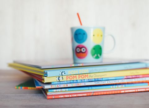 Een stapel kleurrijke kinderboeken ligt op een tafel. Naast de tafel staat een mok met blije gezichten en een rietje.