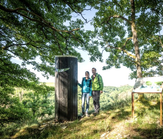 Twee mensen staan naast een houten pilaar in het bos. Rechts op de foto staat een informatiebord. De omgeving is groen en zonnig., © Eifel Tourismus GmbH, Dominik Ketz