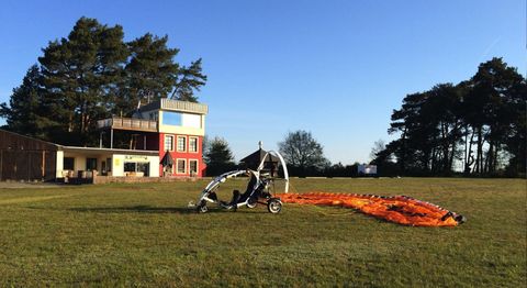 Ein Fluggerät steht auf einer Wiese neben einem modernen Gebäude. Im Hintergrund sind Bäume und ein klarer Himmel zu sehen.