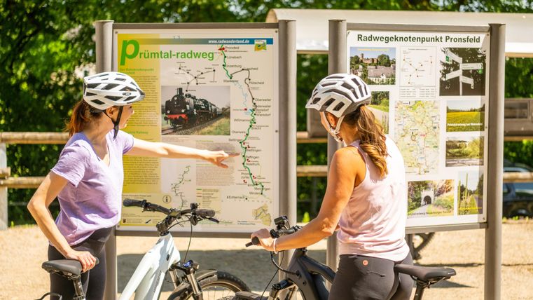 Two female cyclists in helmets are looking at a map of the Prüm Valley cycle path. They are planning their route and seem to be discussing the course of the path.