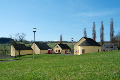 Ein malerisches Dorf mit gelben Häusern und einem großen grünen Rasen. Im Hintergrund sind hohe Bäume und ein klarer blauer Himmel zu sehen.