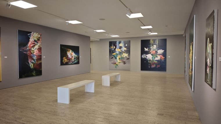 Un espace d'exposition moderne avec plusieurs œuvres d'art colorées sur les murs. Au centre, un banc blanc se trouve sur le sol.