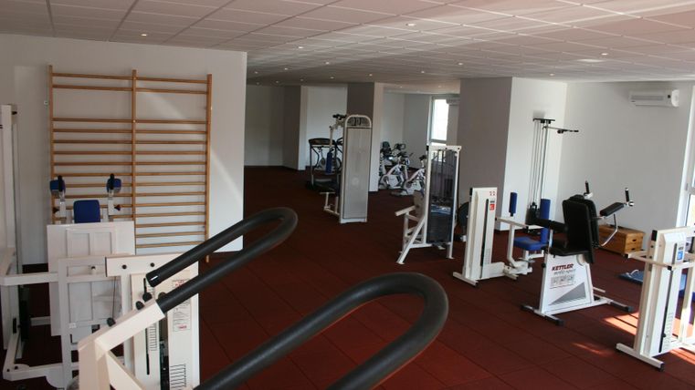 A modern fitness room with various training equipment. The floor is covered with red carpet and large windows provide plenty of daylight.