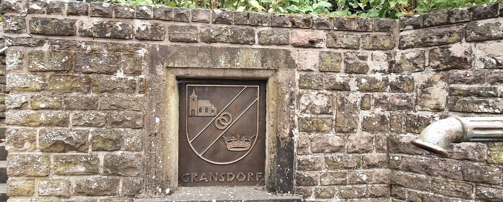 Mur de pierre avec un blason de Gransdorf repr&eacute;sentant une &eacute;glise, deux anneaux et une couronne. A droite, on peut voir un tuyau d'&eacute;vacuation., &copy; TI Bitburger Land S.Wagner