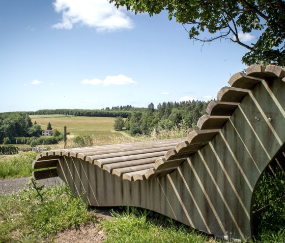 Holzliegebank mit Blick auf gr&uuml;ne Felder und W&auml;lder unter blauem Himmel in Neidenbach., &copy; TI Bitburger Land - Monika Mayer